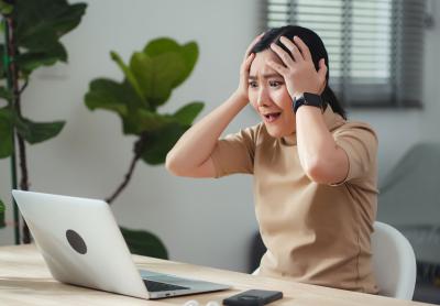 asian woman with hands on her head, expressing frustration and amazement, while looking down at her laptop
