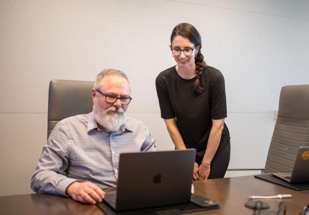 adult man and woman gathered around a conference room table reviewing something on a laptop
