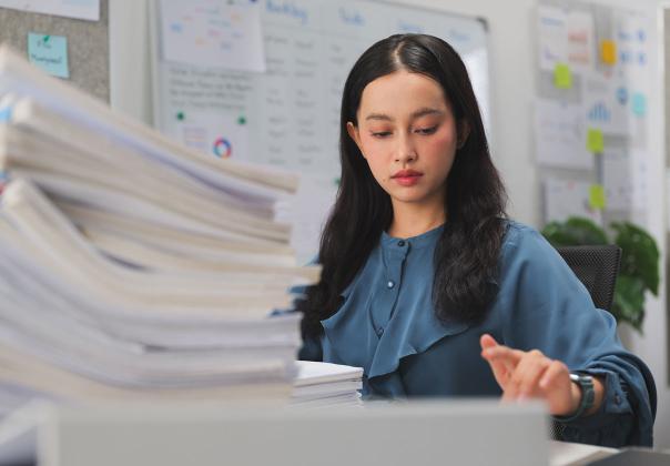 young woman at desk in office with a large stack of papers 