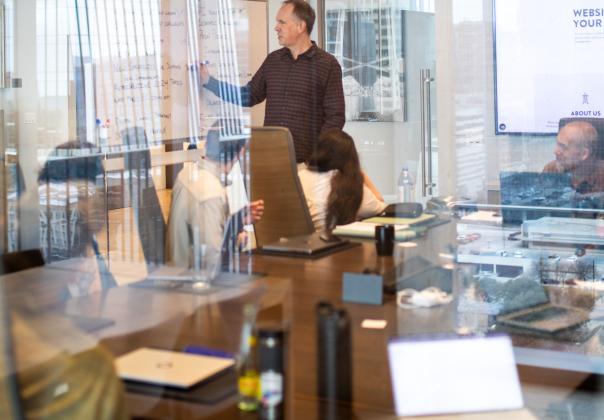 adult male writing on a whiteboard in a conference room, while others seated around the table watch
