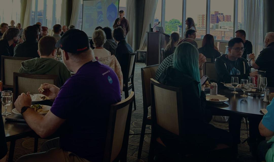 crowd of conference attendees in a large room, sitting at tables and listening to a person speak