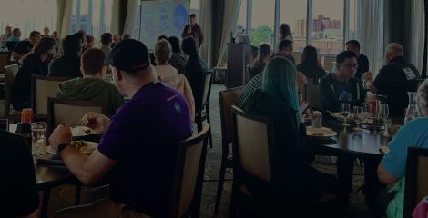 crowd of conference attendees in a large room, sitting at tables and listening to a person speak