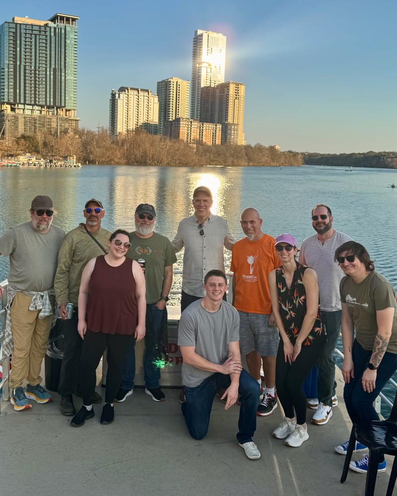 team gathered together on a boat deck, posing with city skyline behind them and the river