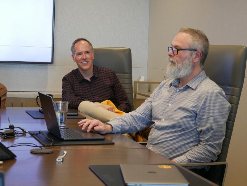 two adult males seated at an office conference room table, chatting with each other