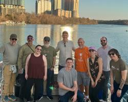 team gathered together on a boat deck, posing with city skyline behind them and the river