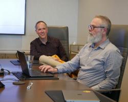 two adult males seated at an office conference room table, chatting with each other