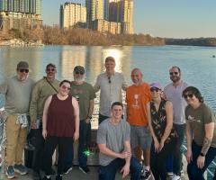 team gathered together on a boat deck, posing with city skyline behind them and the river