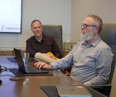 two adult males seated at an office conference room table, chatting with each other