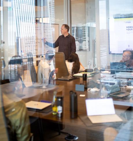 adult male writing on a whiteboard in a conference room, while others seated around the table watch