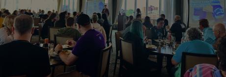 crowd of conference attendees in a large room, sitting at tables and listening to a person speak