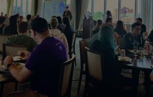 crowd of conference attendees in a large room, sitting at tables and listening to a person speak